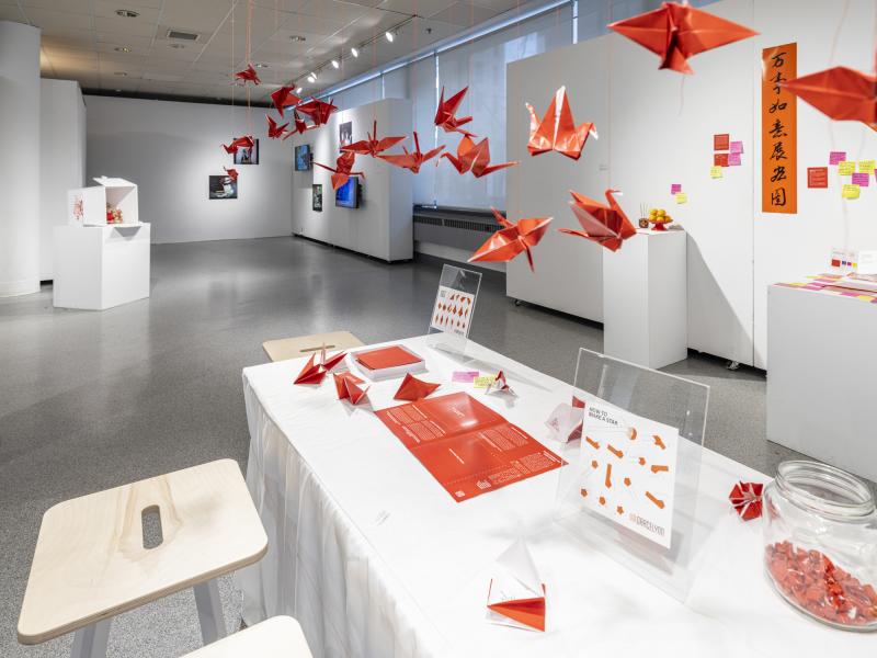 installation image of hanging origami birds, items displayed on a table, sculptures, and video monitors