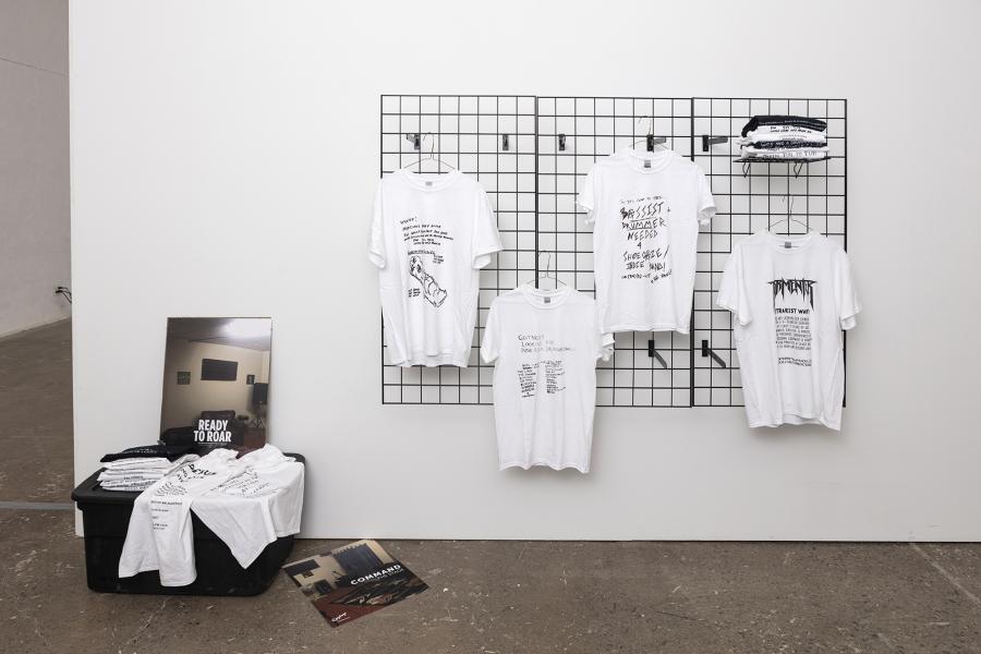 Image of t-shirts hanging on a black grid with posters and a black plastic bin with t-shirts on the ground to the left