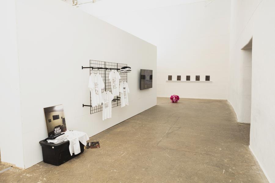 installation image of multiple artworks including hanging t-shirts, video monitor, floor sculpture, and ceramics