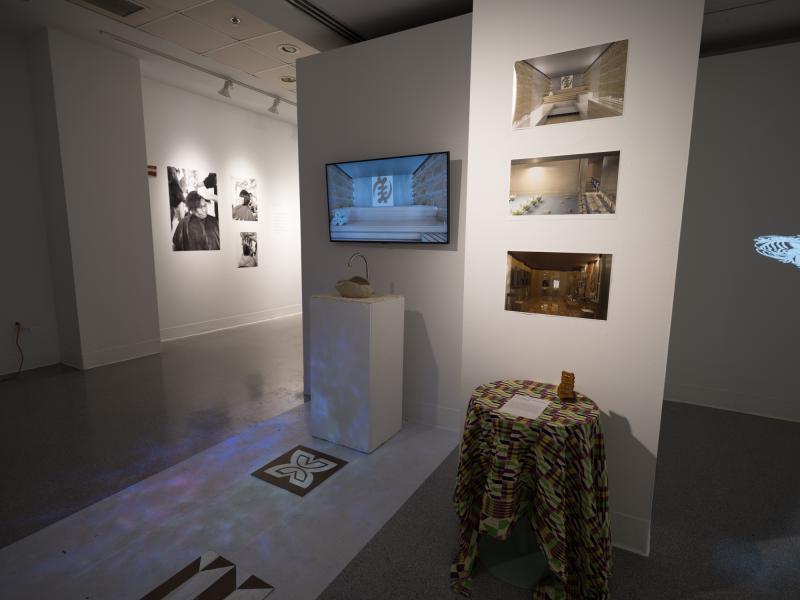 installation image of a dark space with photographs, monitor, side table and floor projection
