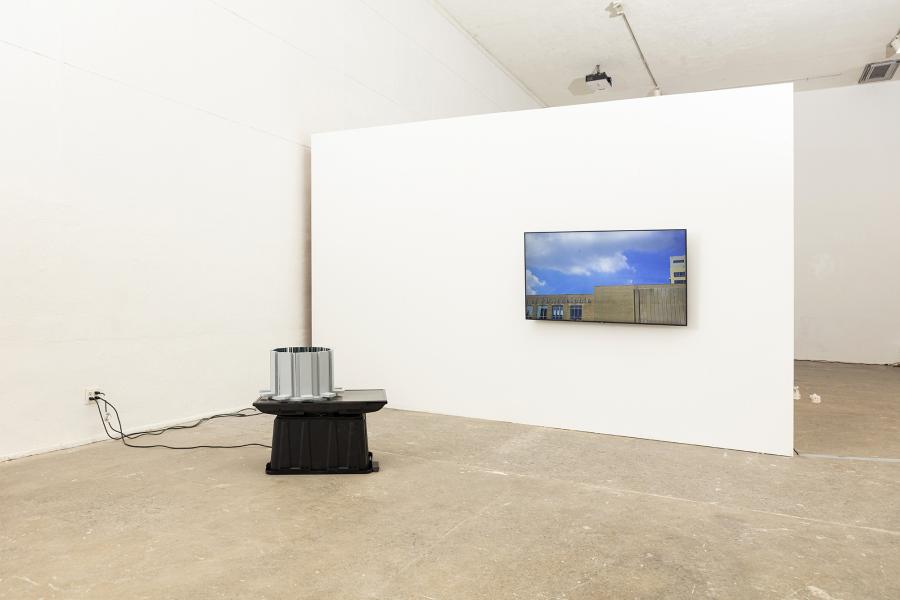 image of a floor sculpture with monitor and plastic bin to the left and wall mounted monitor displaying helicopters to the right