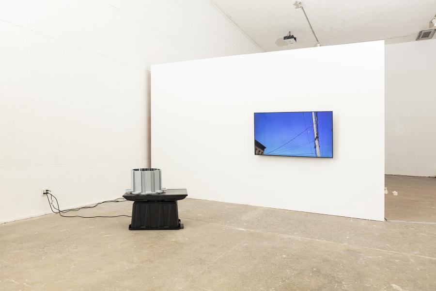 image of a floor sculpture with monitor and plastic bin to the left and wall mounted monitor displaying helicopters to the right