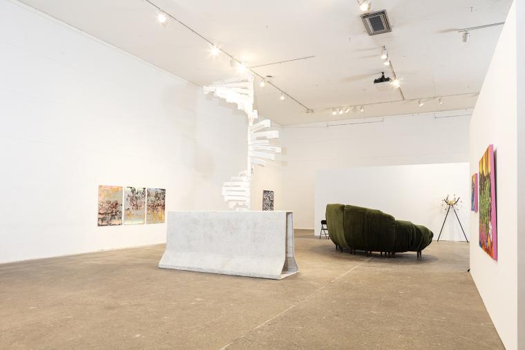 Gallery space with paintings on walls, black leather couch, white traffic barrier and white spiral staircase