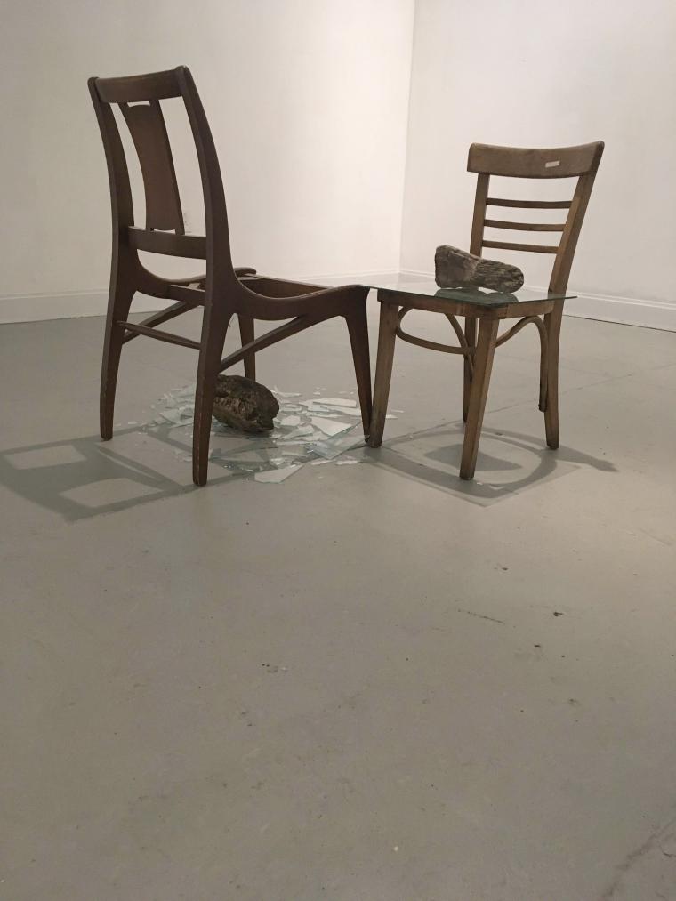 sculpture using two wooden chairs, rocks, a pane of glass, and broken glass on the floor