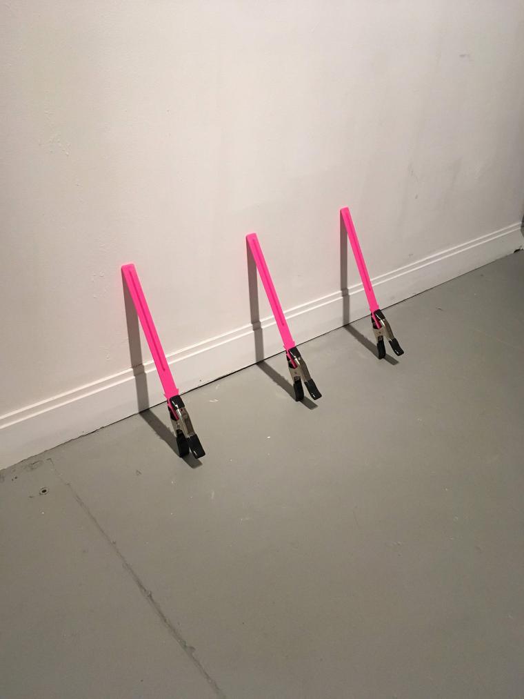 three small identical sculptures leaning against the wall using clamps and bright pink plastic stakes